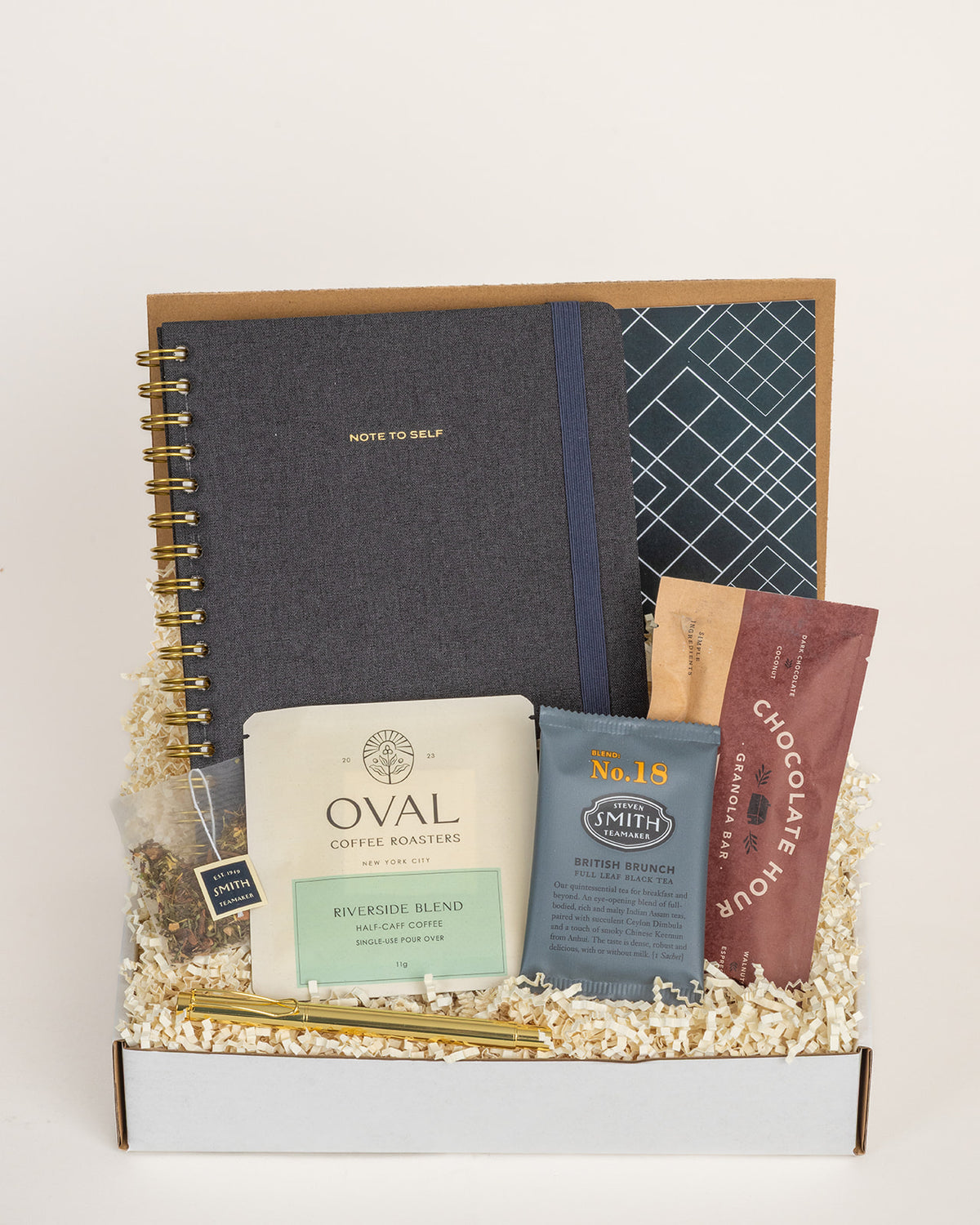 Gift box with a charcoal linen notebook, gold pen, pour-over coffee, black tea, and a granola bar on paper filler.
