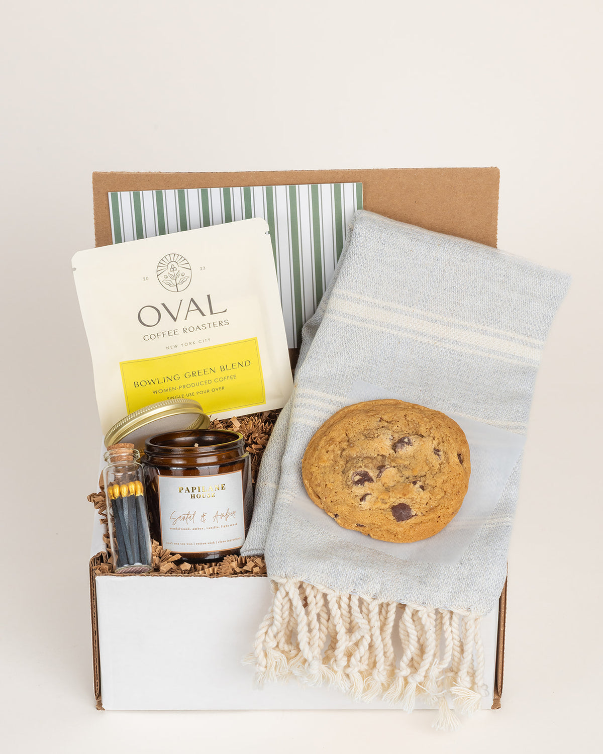 Gift box with a Turkish towel, chocolate chip cookie, Oval coffee packet, amber candle, and matches in a glass vial.