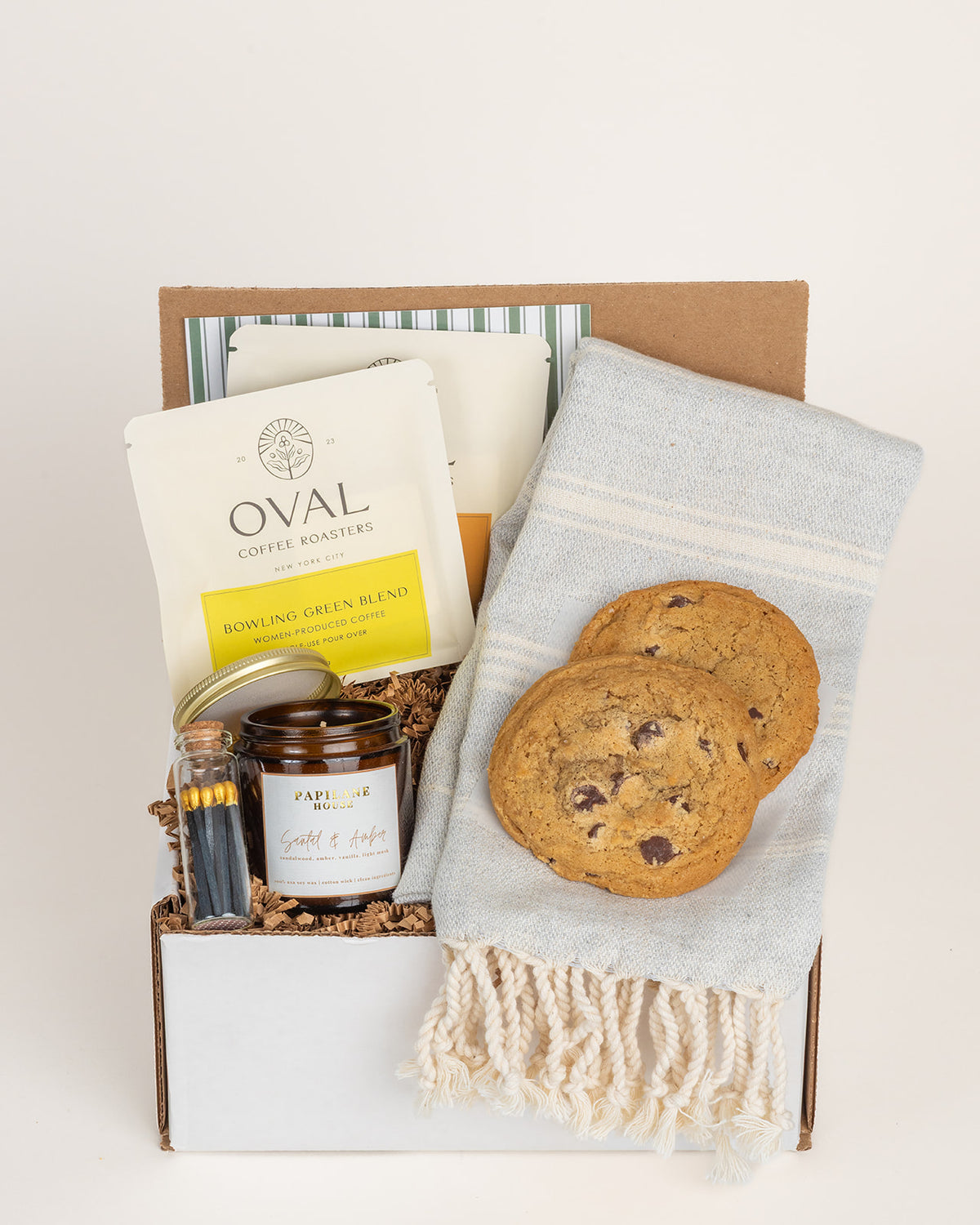 Gift box with two chocolate chip cookies, a blue Turkish towel, Oval coffee packets, a scented candle, and matches.