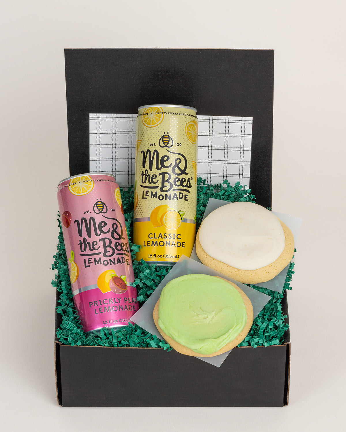 Gift box with two Me & the Bees lemonade cans and two frosted citrus cookies nestled in green paper shred.