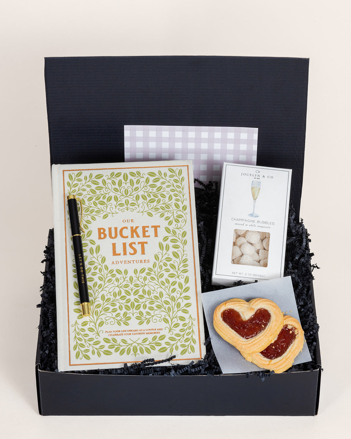 Black gift box containing a bucket list book, motivational pen, champagne gummies, and heart-shaped jam cookies.
