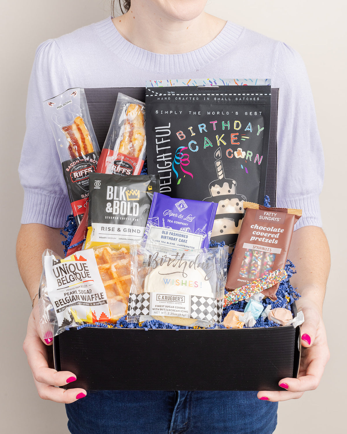 Person holding a black gift box filled with birthday treats, including popcorn, Belgian waffles, cookies, and chocolate pretzels.