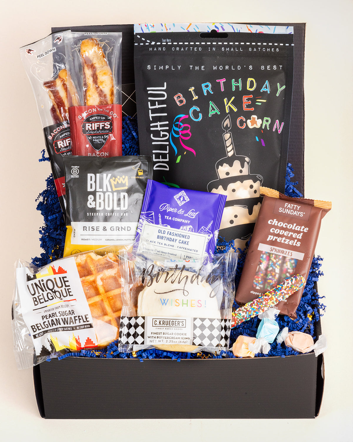 Birthday gift box filled with popcorn, Belgian waffle, cookies, coffee, tea, chocolate pretzels, and bacon on blue paper filler.