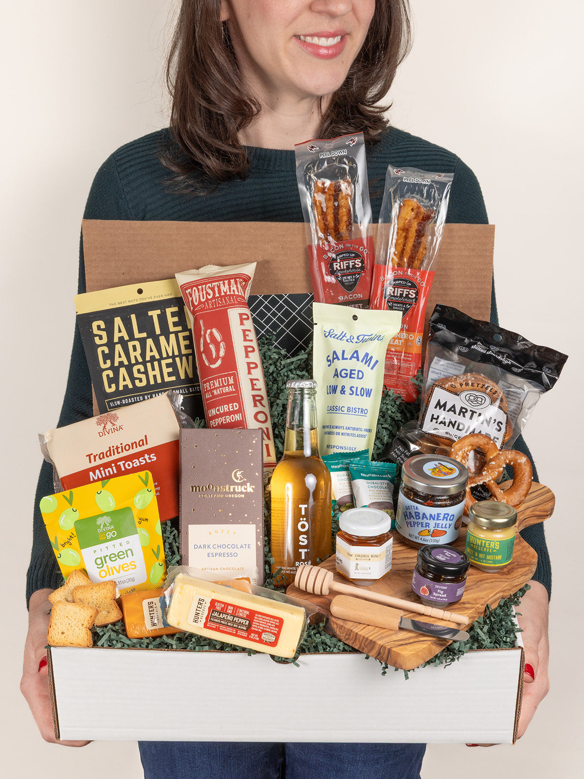 Person holding a gift box filled with assorted cheeses, meats, snacks, chocolates, and a non-alcoholic beverage on an olive wood board.