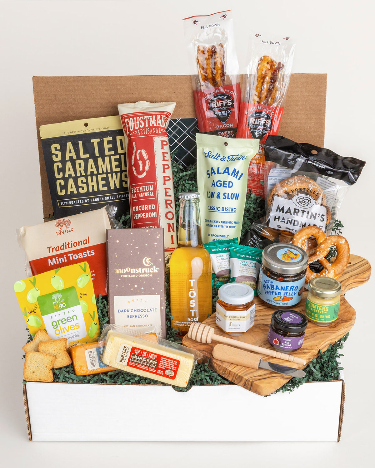 Charcuterie-in-a-Box gift set with assorted cheeses, meats, snacks, and a wooden board on a green paper-filled base.