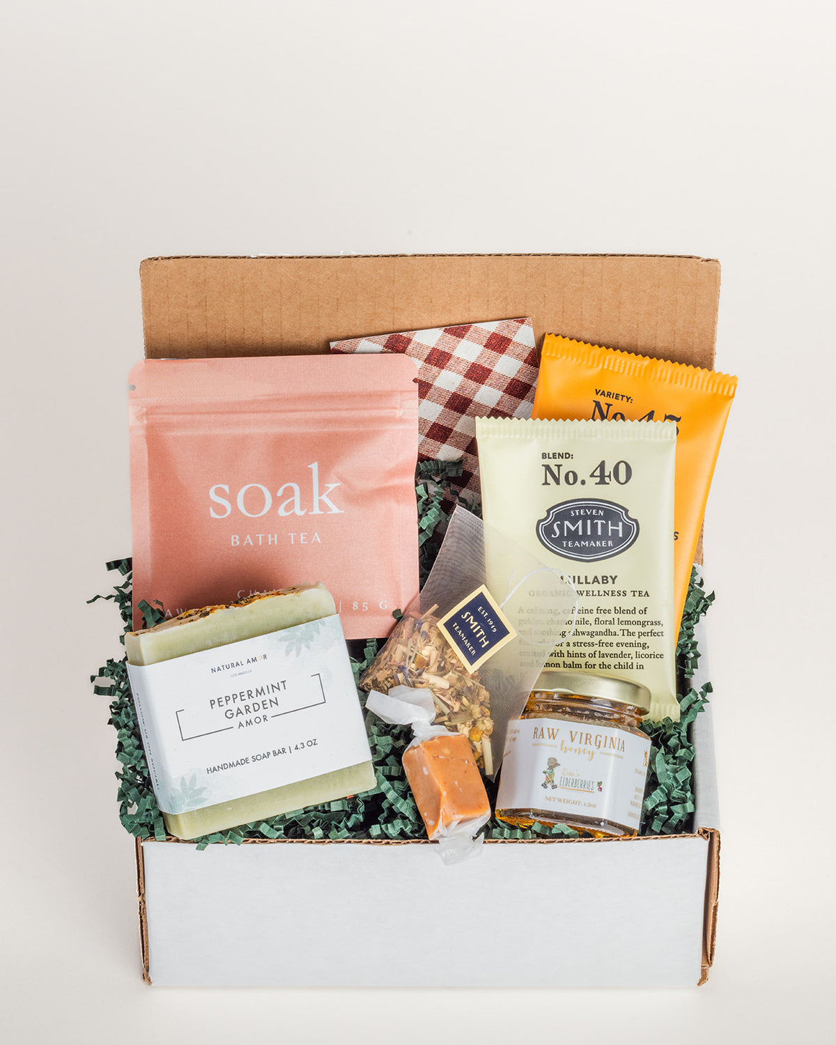 Care package in a cardboard box with bath tea, peppermint soap, elderberry honey, tea sachets, and a caramel on green filler.