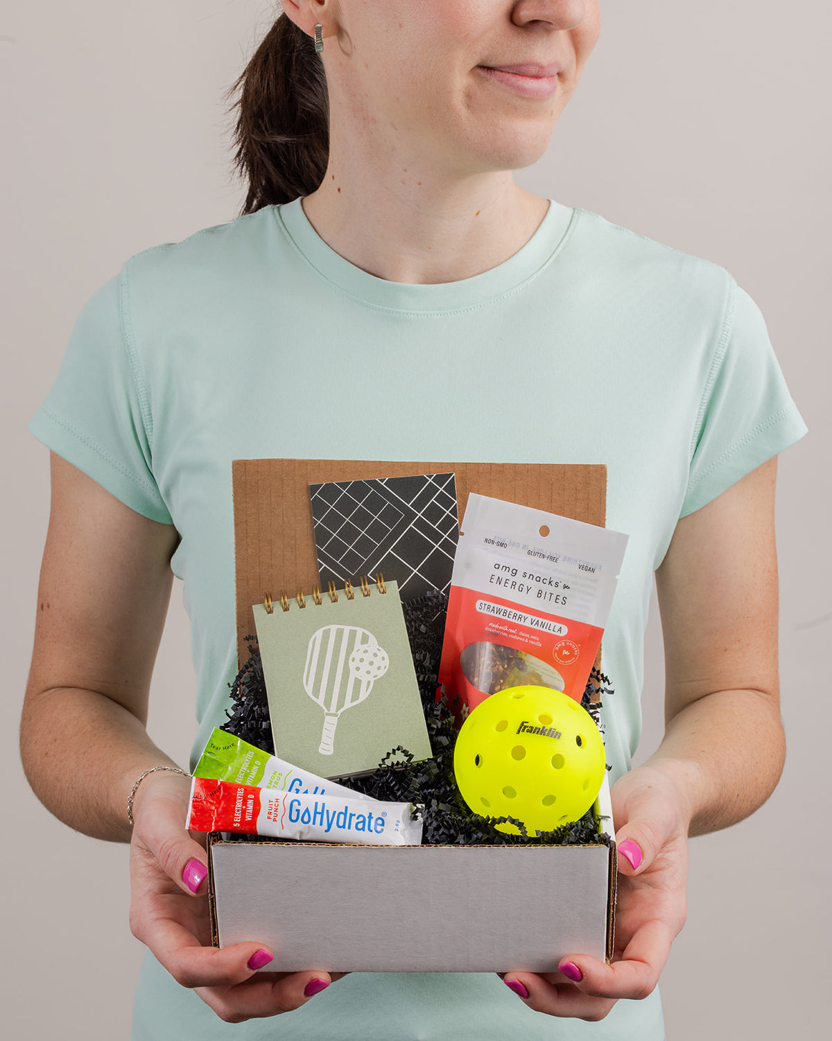 Person holding a gift box with a pickleball, energy bites, electrolytes, and a pickleball-themed jotter pad.