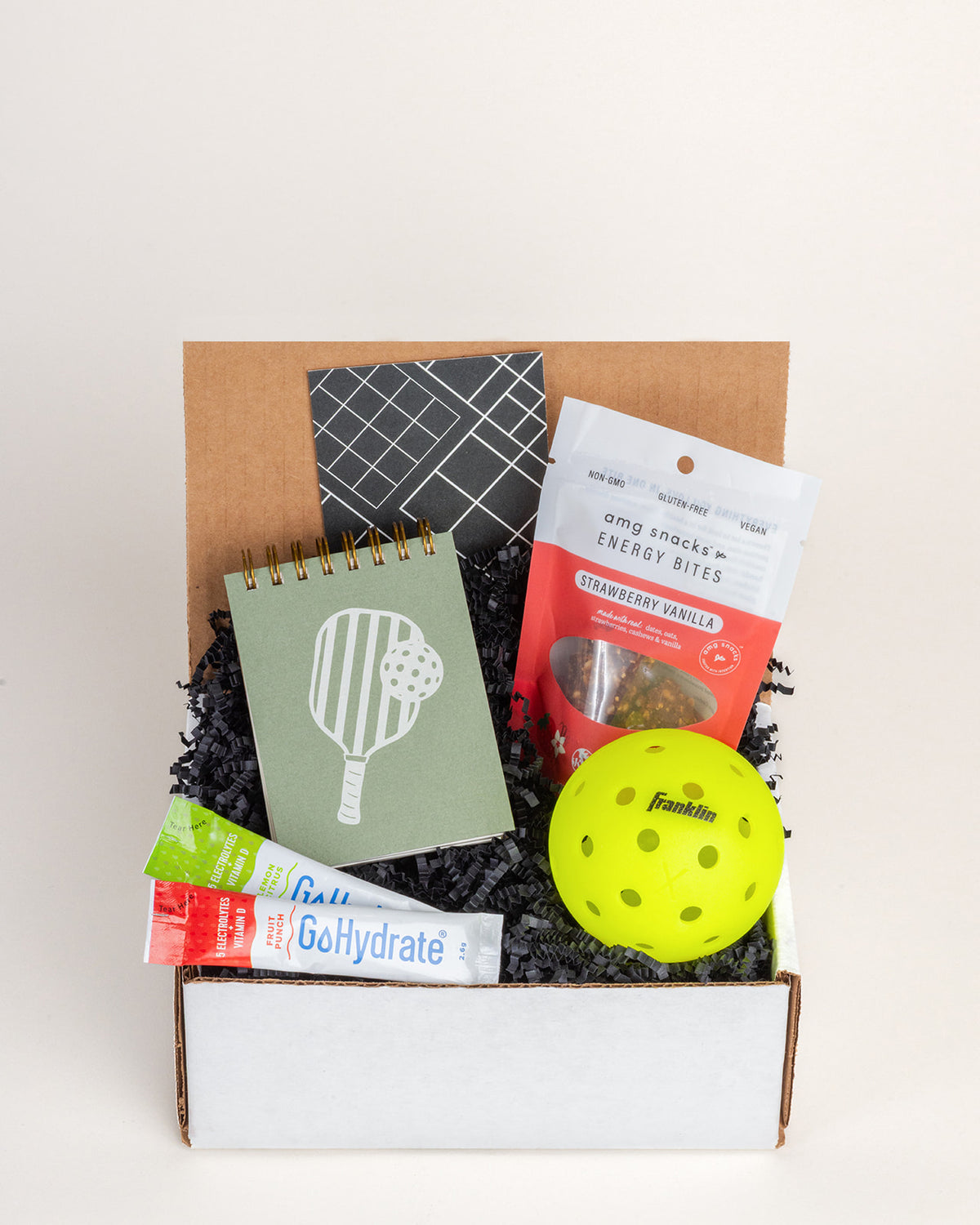 Pickleball gift box with a green jotter pad, yellow ball, energy bites, and electrolyte packets in black crinkle paper.