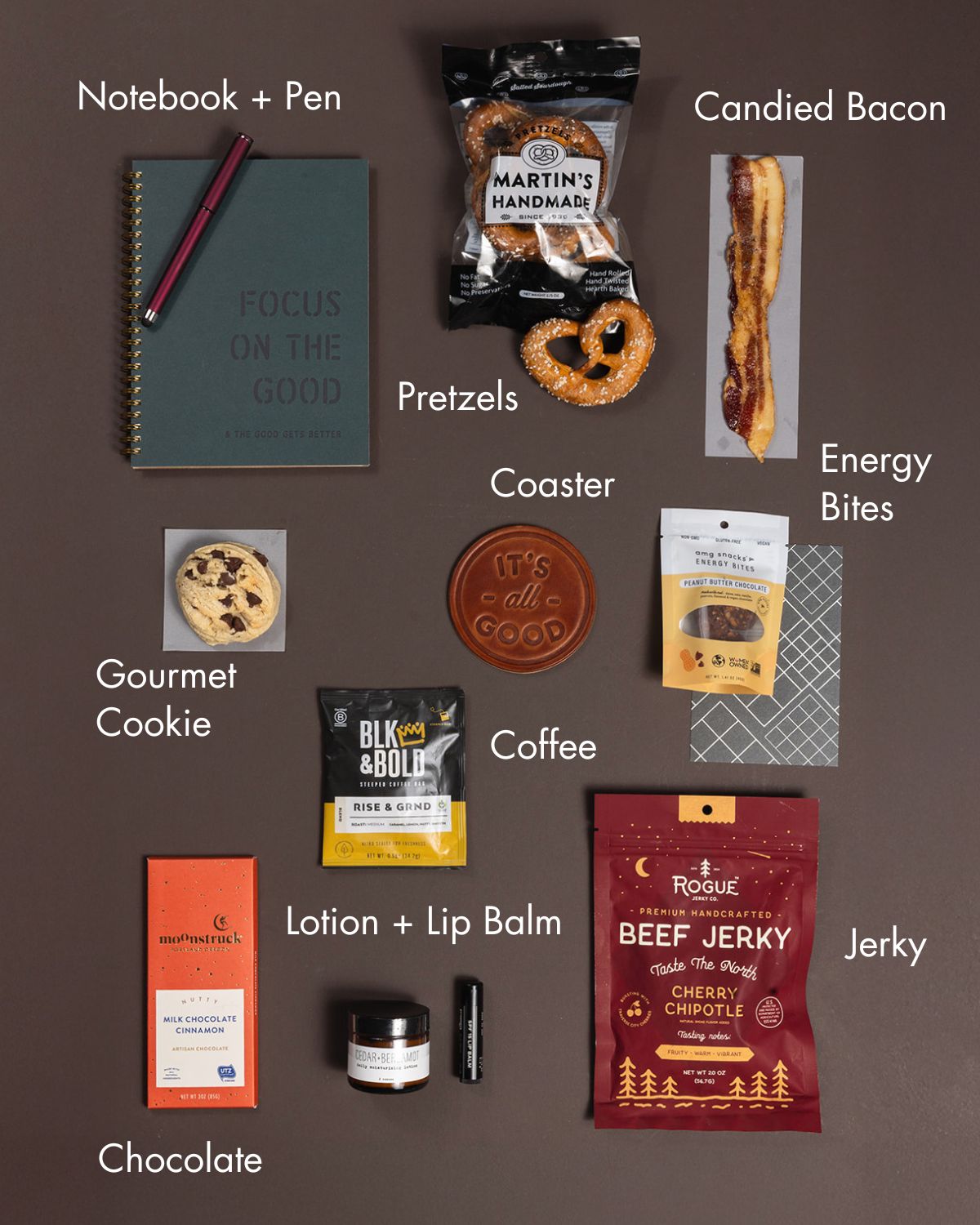 Focus On The Good gift box with a notebook, pen, leather coaster, snacks, coffee, chocolate, and skincare on a brown background.