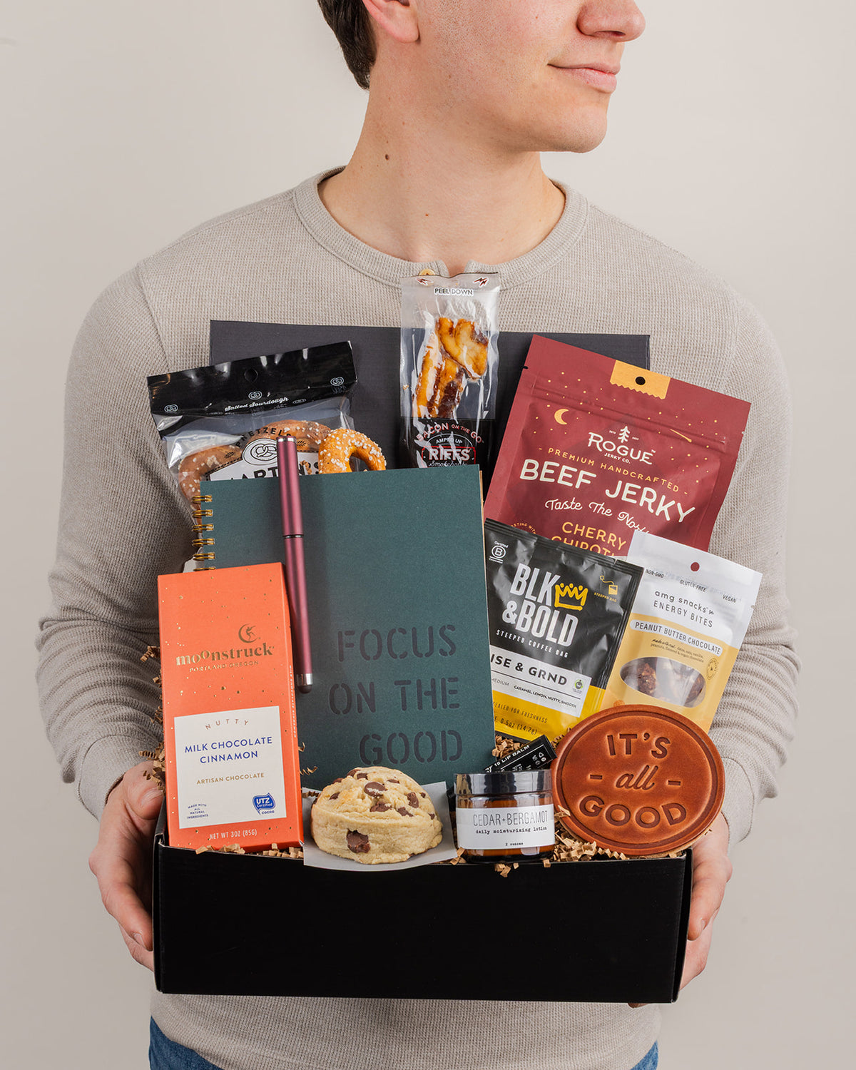 Person holding a Focus On The Good gift box filled with snacks, a notebook, coffee, and skincare essentials.