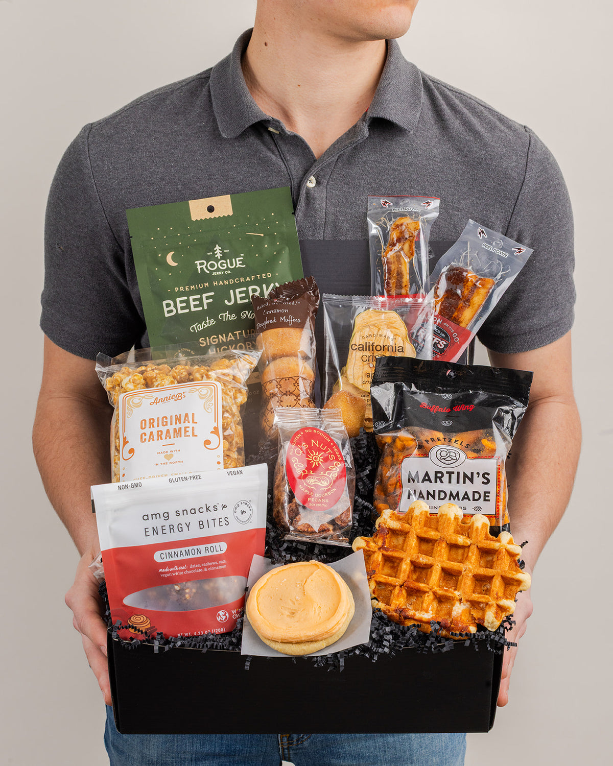 Person holding a black gift box filled with assorted snacks including beef jerky, caramel popcorn, a waffle, and cookies.