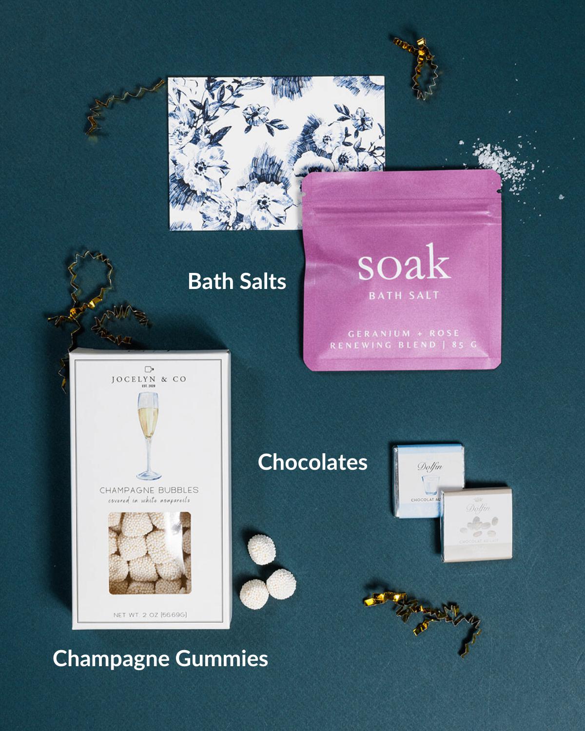 Gift set with pink bath salts, champagne gummies in a white box, two Belgian chocolates, and a floral note on teal.