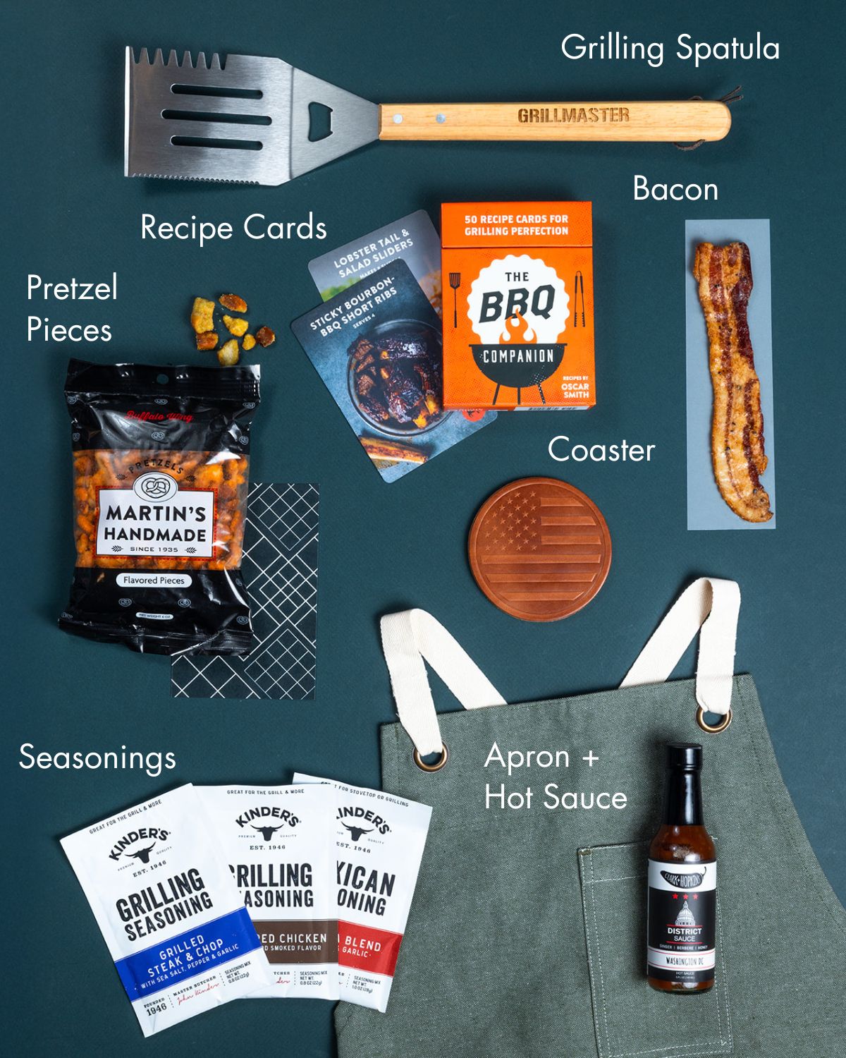 Grilling gift set with a metal spatula, BBQ recipe cards, seasoning packets, hot sauce, pretzel snacks, and a green apron.