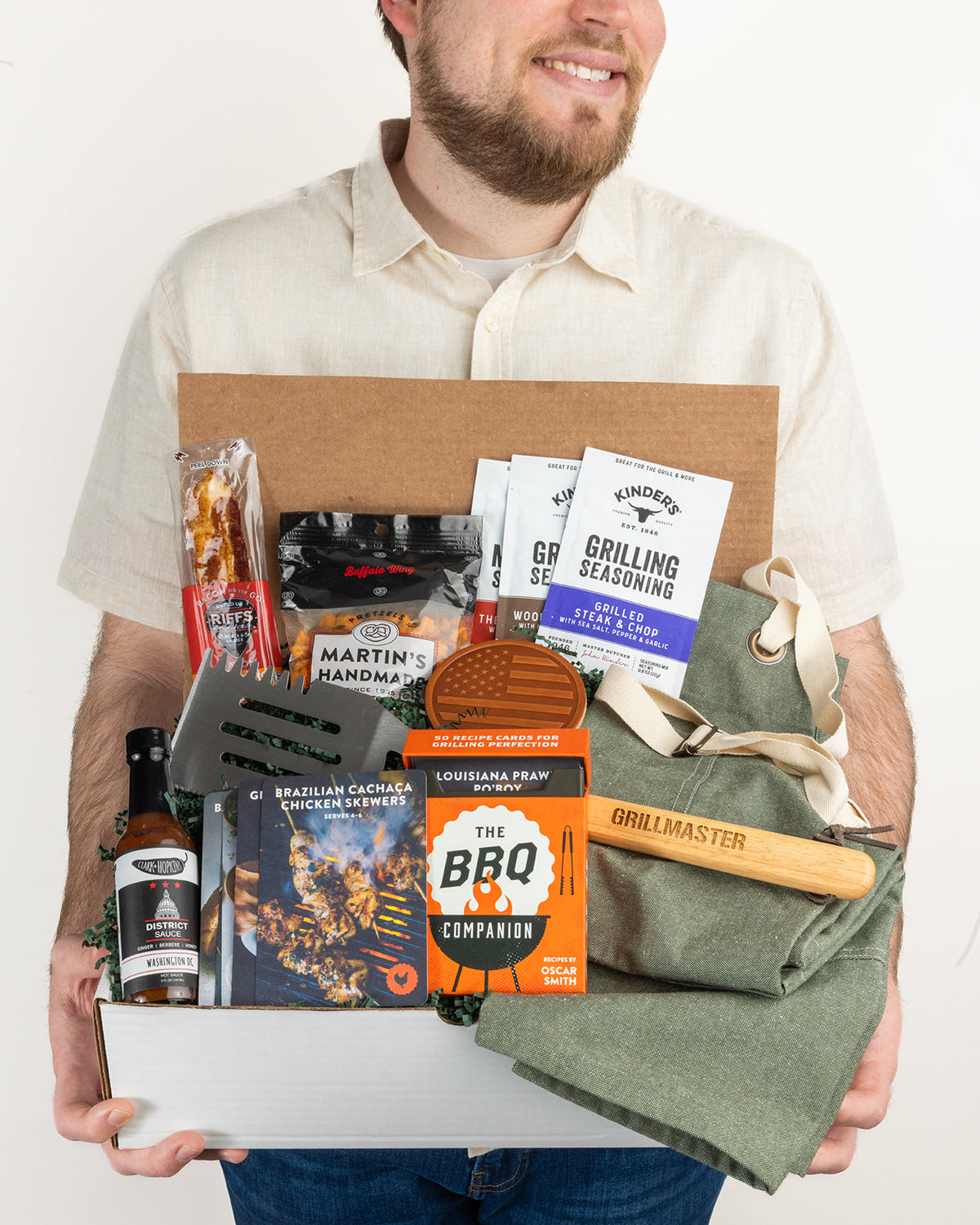 Man holding a grill gift box with a green apron, spatula, hot sauce, seasoning packets, recipe cards, and snacks.