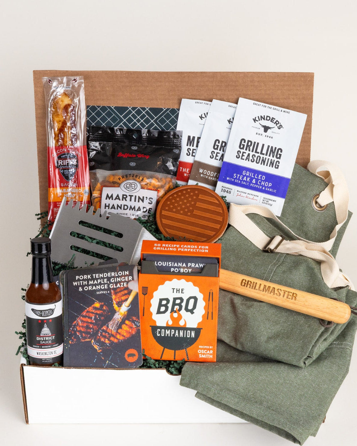 Grilling gift box with olive apron, metal spatula, hot sauce, seasoning packets, recipe cards, and snacks in a cardboard crate.