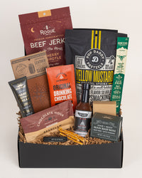 Rugged gift box featuring beef jerky, leather koozie, hot sauce, snacks, and grooming essentials in a black box with paper filler.