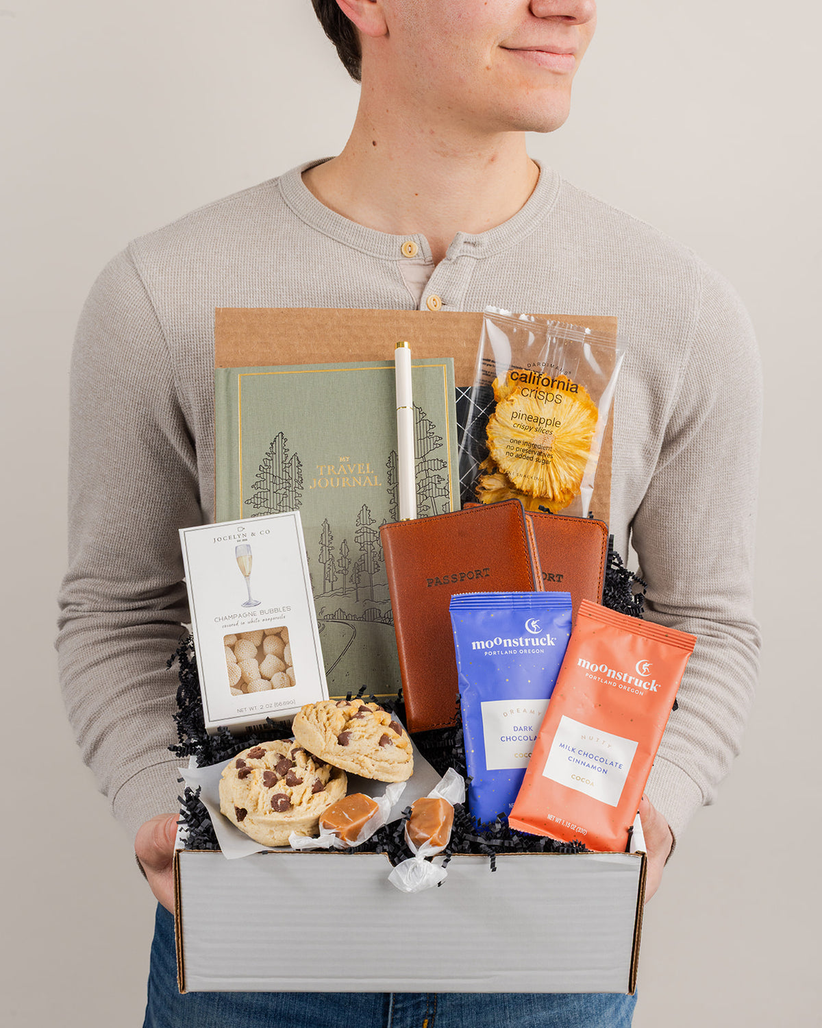 Gift box for couples featuring a sage green travel journal, two brown leather passport covers, gourmet snacks, and sweets.