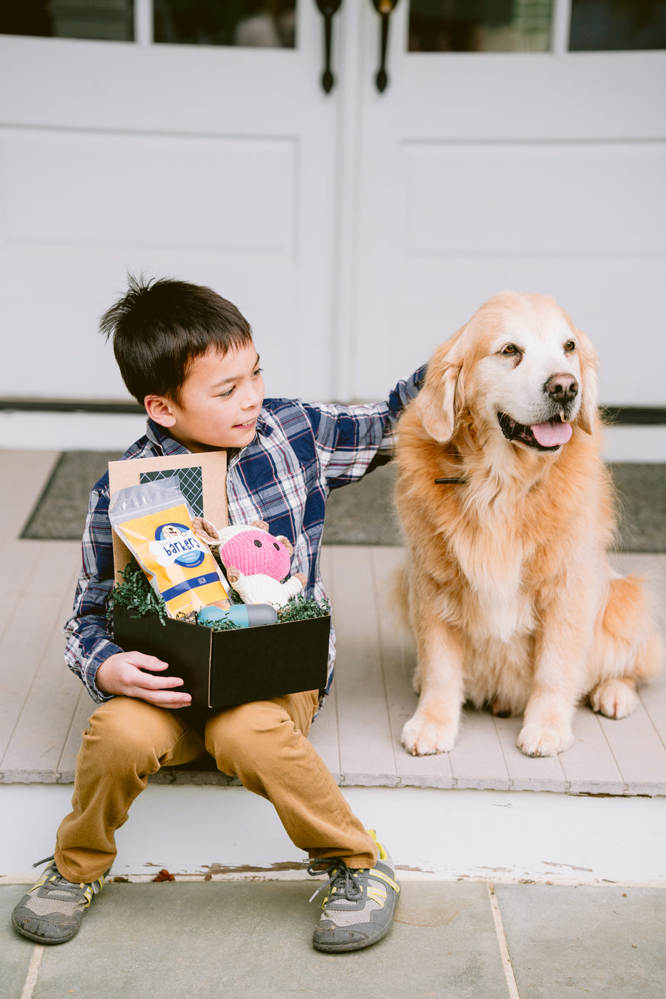 A boy in a plaid shirt sits on a porch holding a black gift box filled with dog treats, a squeaky toy, and a flashlight.