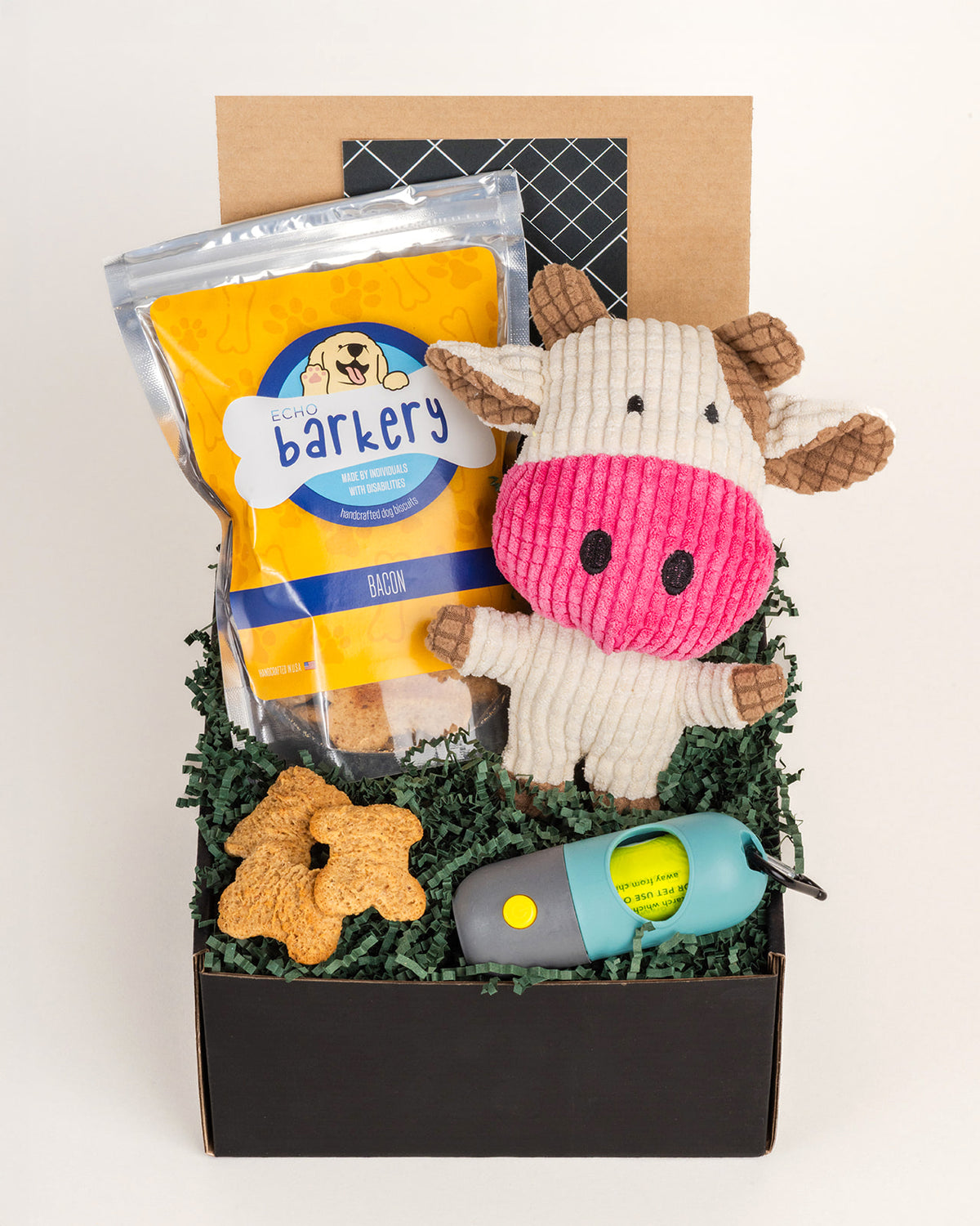 Gift box with Echo Barkery bacon dog treats, a plush cow toy, and a teal flashlight bag dispenser on green crinkle paper.
