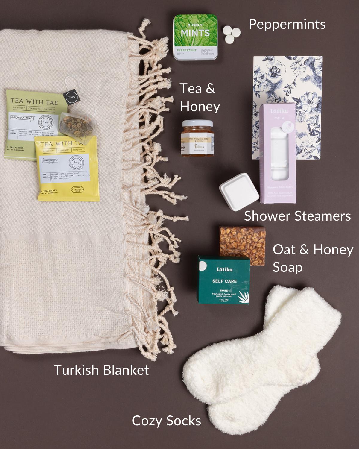 Get Well care package featuring a cream Turkish blanket, cozy white socks, herbal tea, honey, mints, shower steamers, and oat soap.