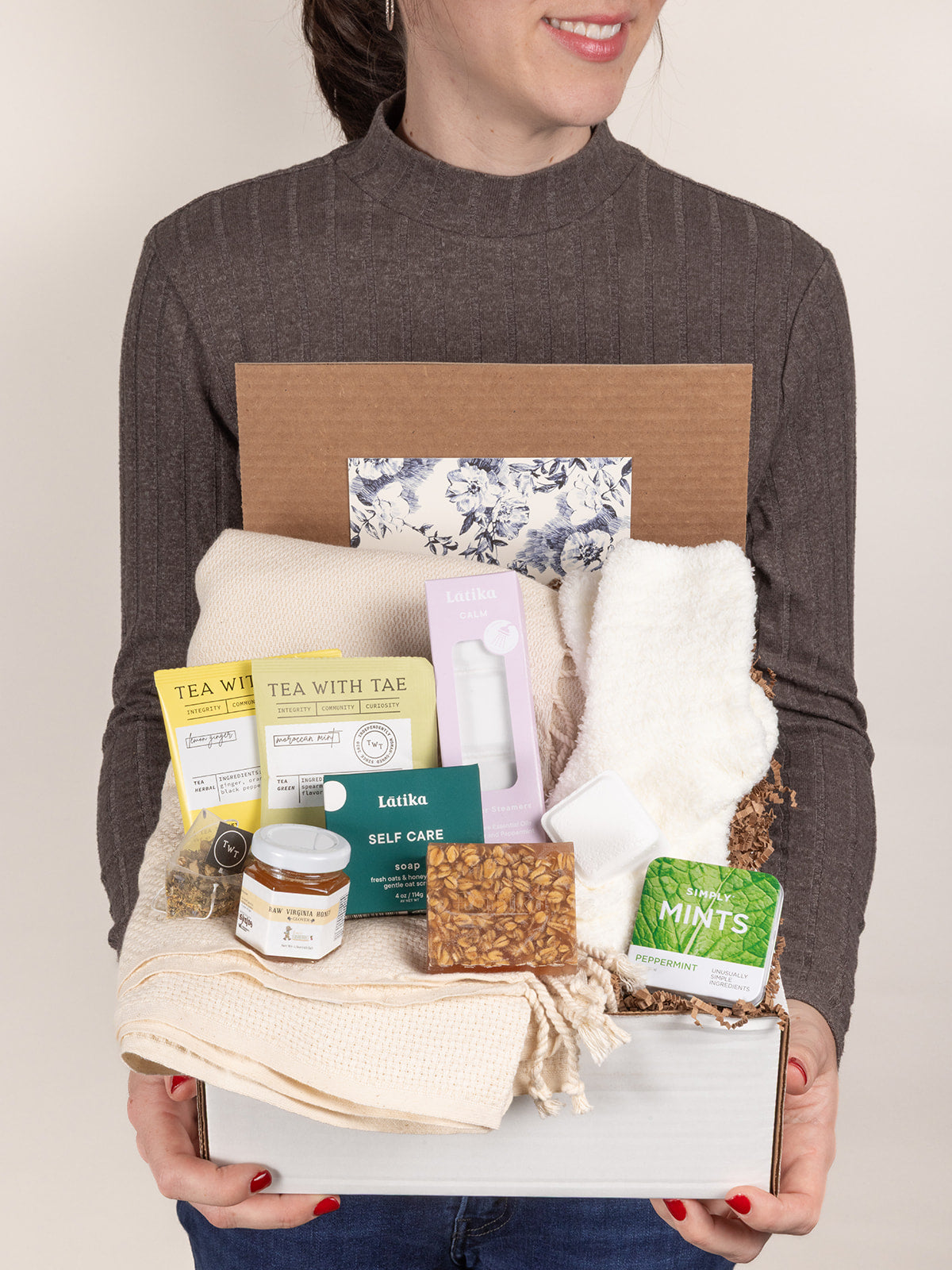 Person holding a get well gift box with a cozy blanket, tea, honey, soap, shower steamers, fuzzy socks, and mints.