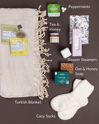 Get Well care package featuring a cream Turkish blanket, cozy white socks, herbal tea, honey, mints, shower steamers, and oat soap.