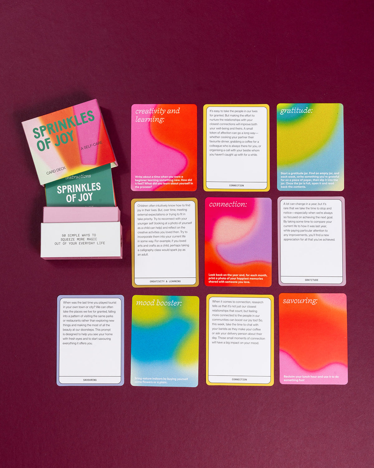 Sprinkles of Joy self-care card deck with colorful, gradient-designed prompt cards laid out on a deep burgundy background.