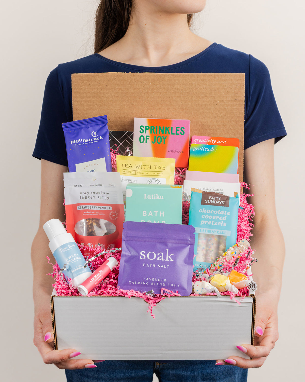 Person holding a Happy Box gift set filled with colorful self-care items, snacks, bath products, and pink paper filler.