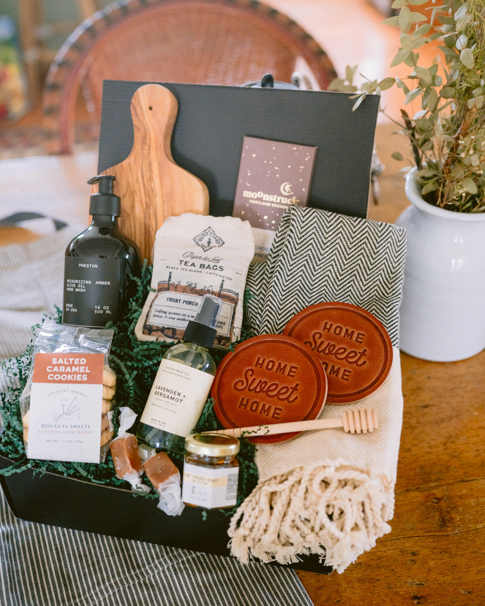 Hearth & Home gift box featuring an olive wood board, leather coasters, hand soap, tea, cookies, and a herringbone throw blanket.
