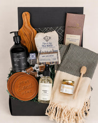 Hearth & Home gift box with olive wood board, tea, leather coasters, hand soap, chocolate, and a black and white patterned towel.