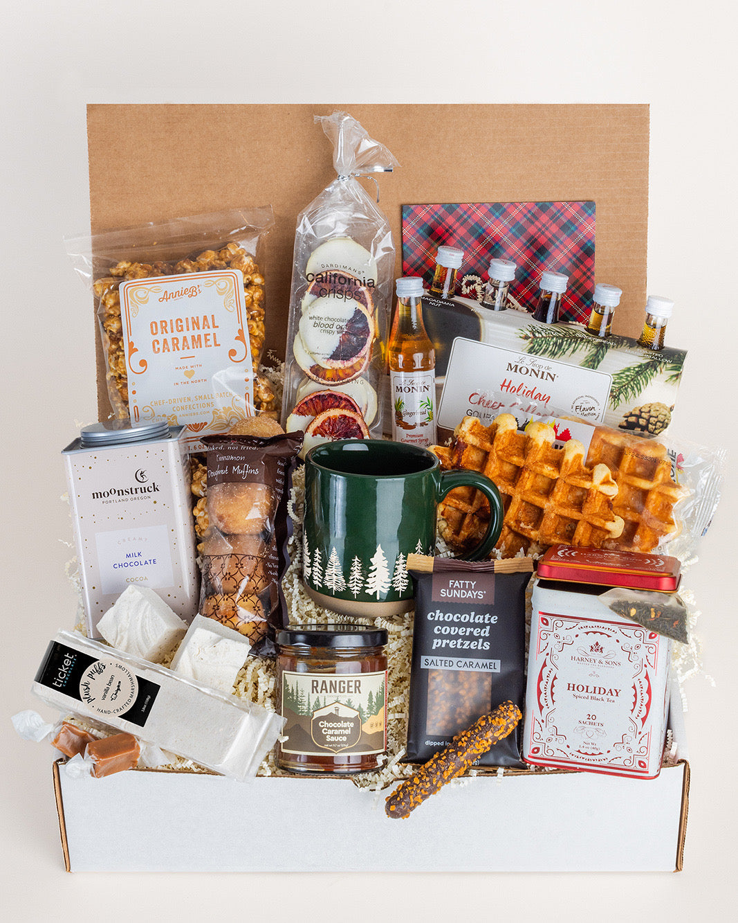 Holiday gift box featuring a green forest-print mug, assorted gourmet snacks, Monin syrup set, and holiday tea in a cardboard crate.