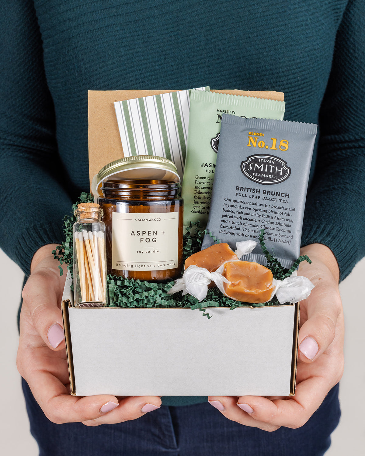 Gift box containing an Aspen and Fog soy candle, Smith tea sachets, matches in a glass vial, and caramels on green paper filler.
