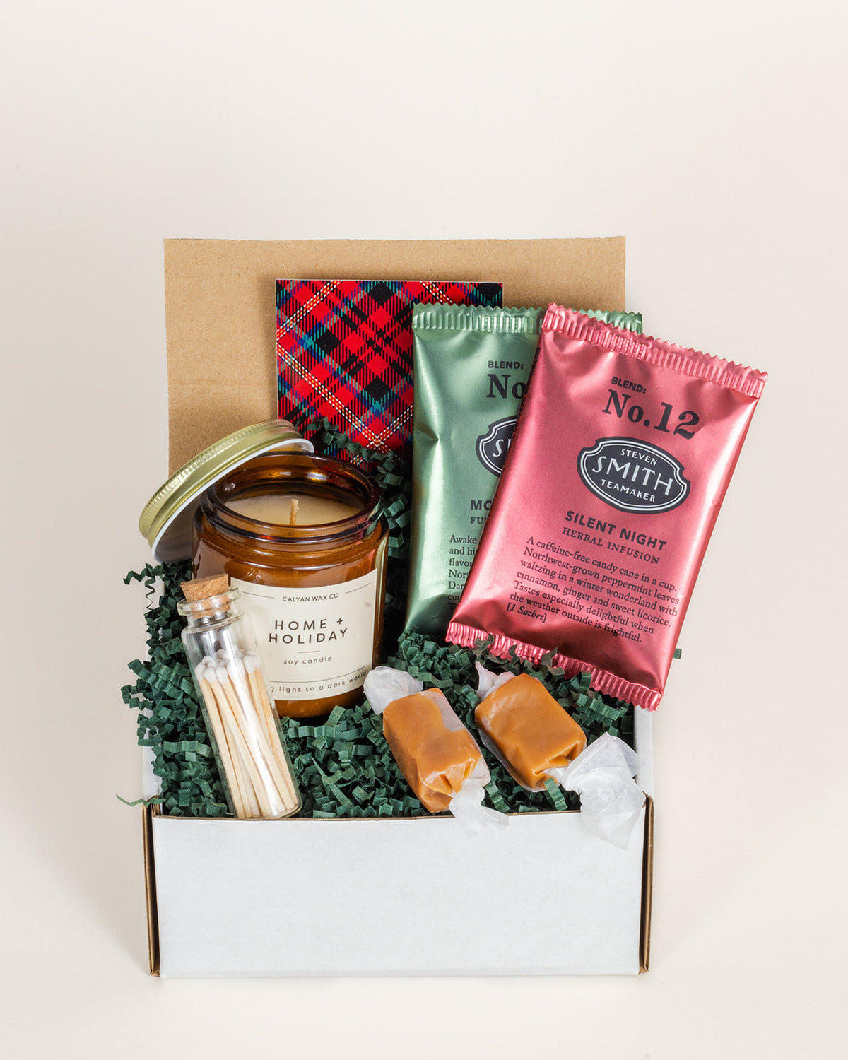 Holiday gift box with a soy candle, tea sachets, matches in a glass vial, and sea salt caramels on green paper filler.