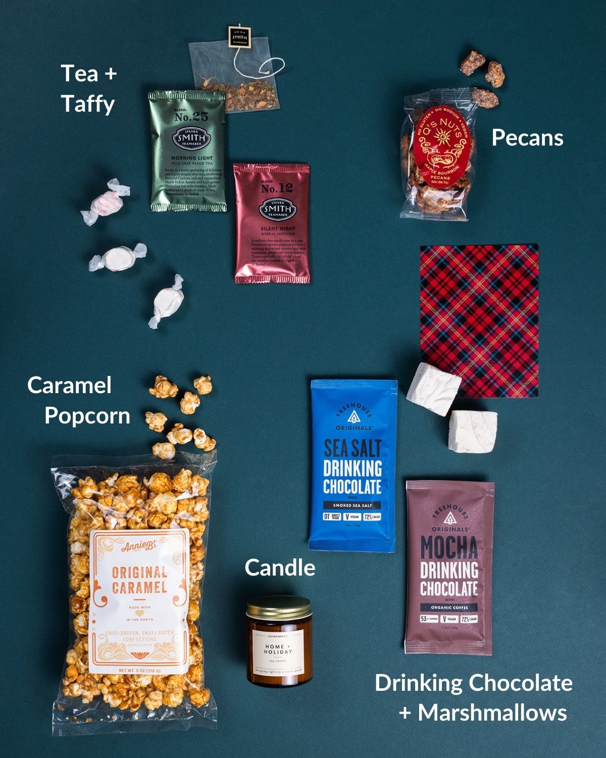 Holiday gift set featuring caramel popcorn, drinking chocolates, tea, candied pecans, marshmallows, and a candle on dark green.