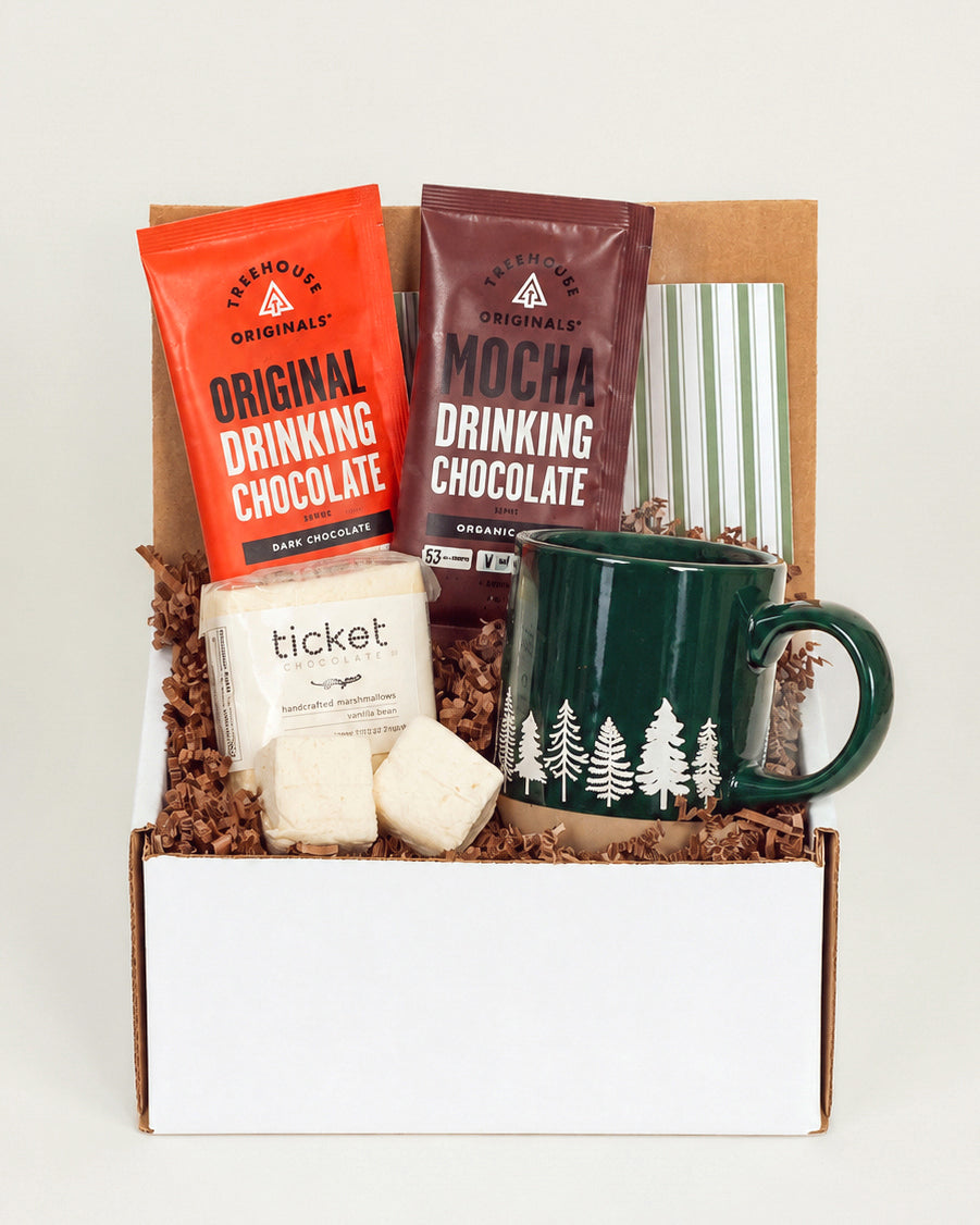 Gift box with a green stoneware mug, drinking chocolate packets, and vanilla bean marshmallows on brown paper filler.