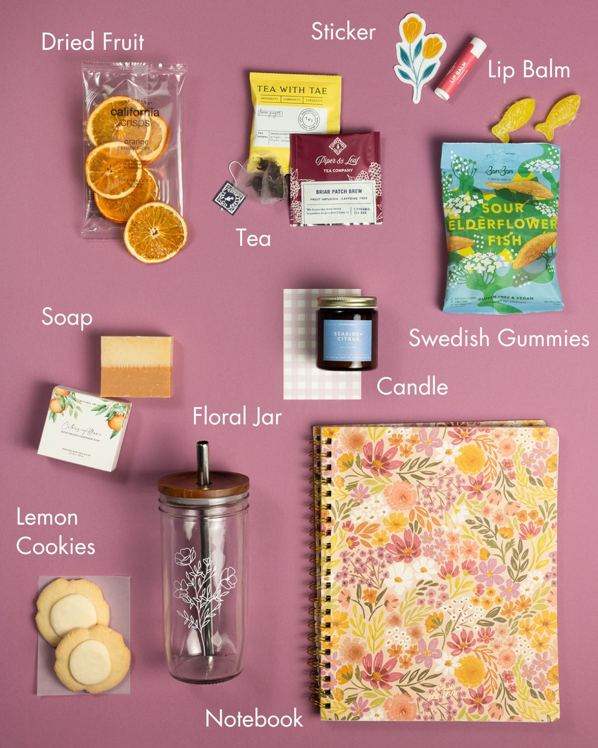 In Full Bloom gift set with floral notebook, glass jar, citrus snacks, tea, candle, soap, and lip balm on a pink background.