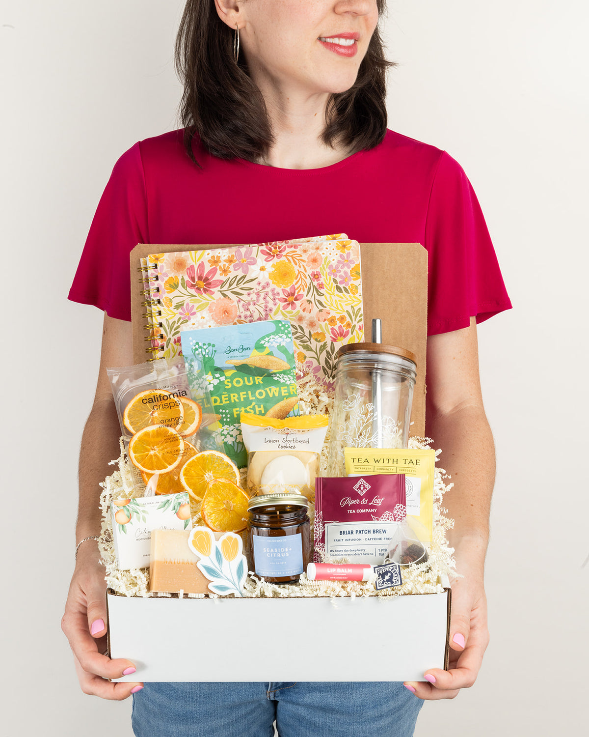 Person holding a gift box filled with floral stationery, citrus snacks, lemon cookies, tea, a glass tumbler, and a candle.