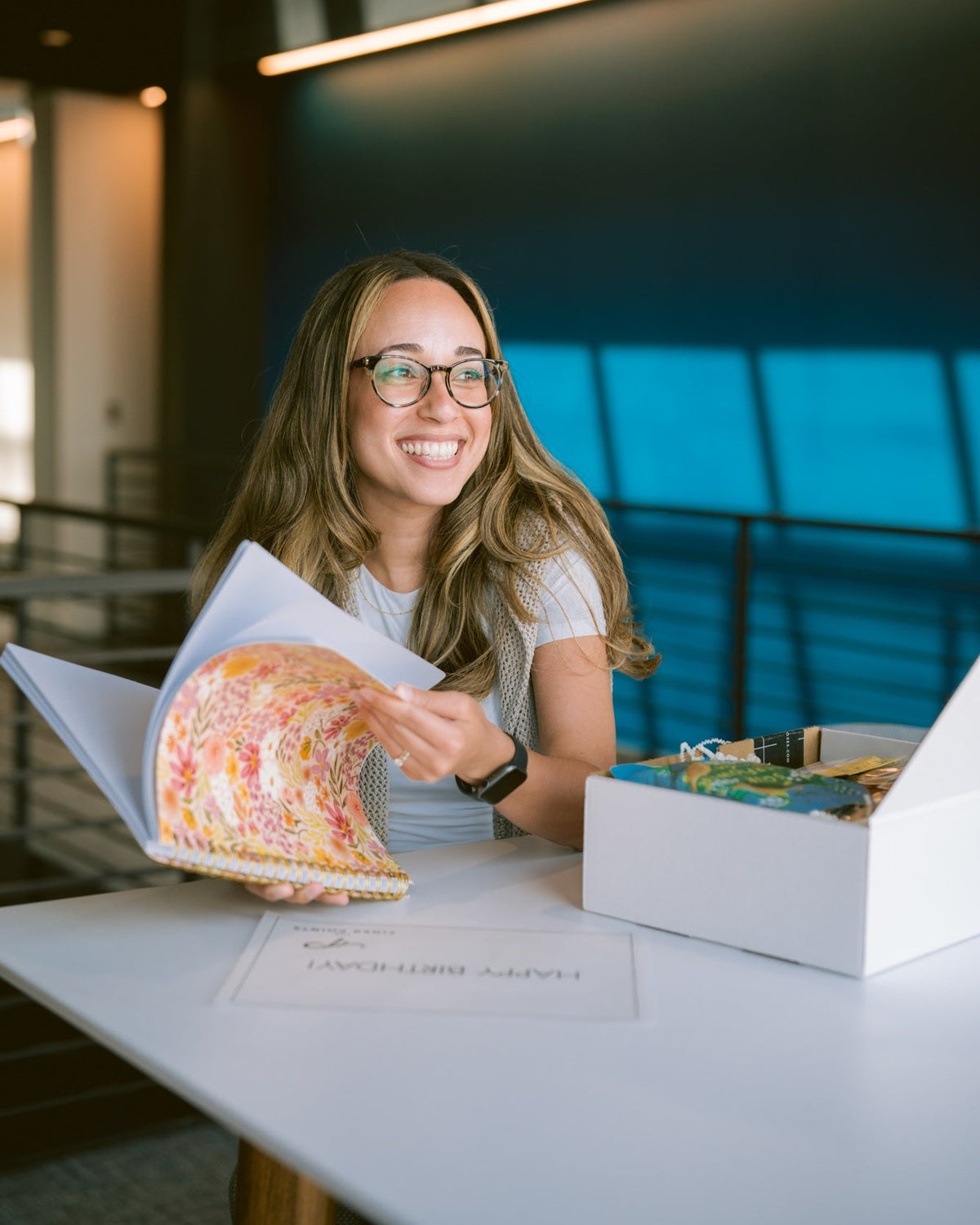 Smiling woman opening an In Full Bloom gift box featuring a floral notebook, snacks, and self-care items on a white table.