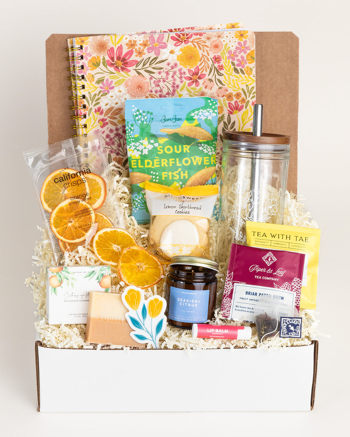 Gift box with floral notebook, glass jar, citrus crisps, lemon cookies, tea, candle, soap, and lip balm on paper shred.