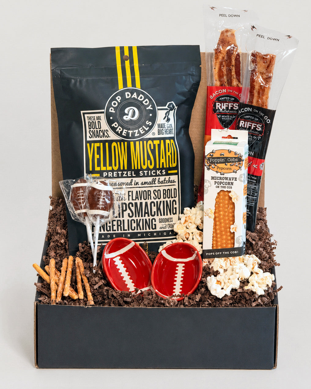 Football-themed snack gift box featuring Pop Daddy pretzels, candied bacon, popcorn on the cob, lollipops, and two red dip bowls.