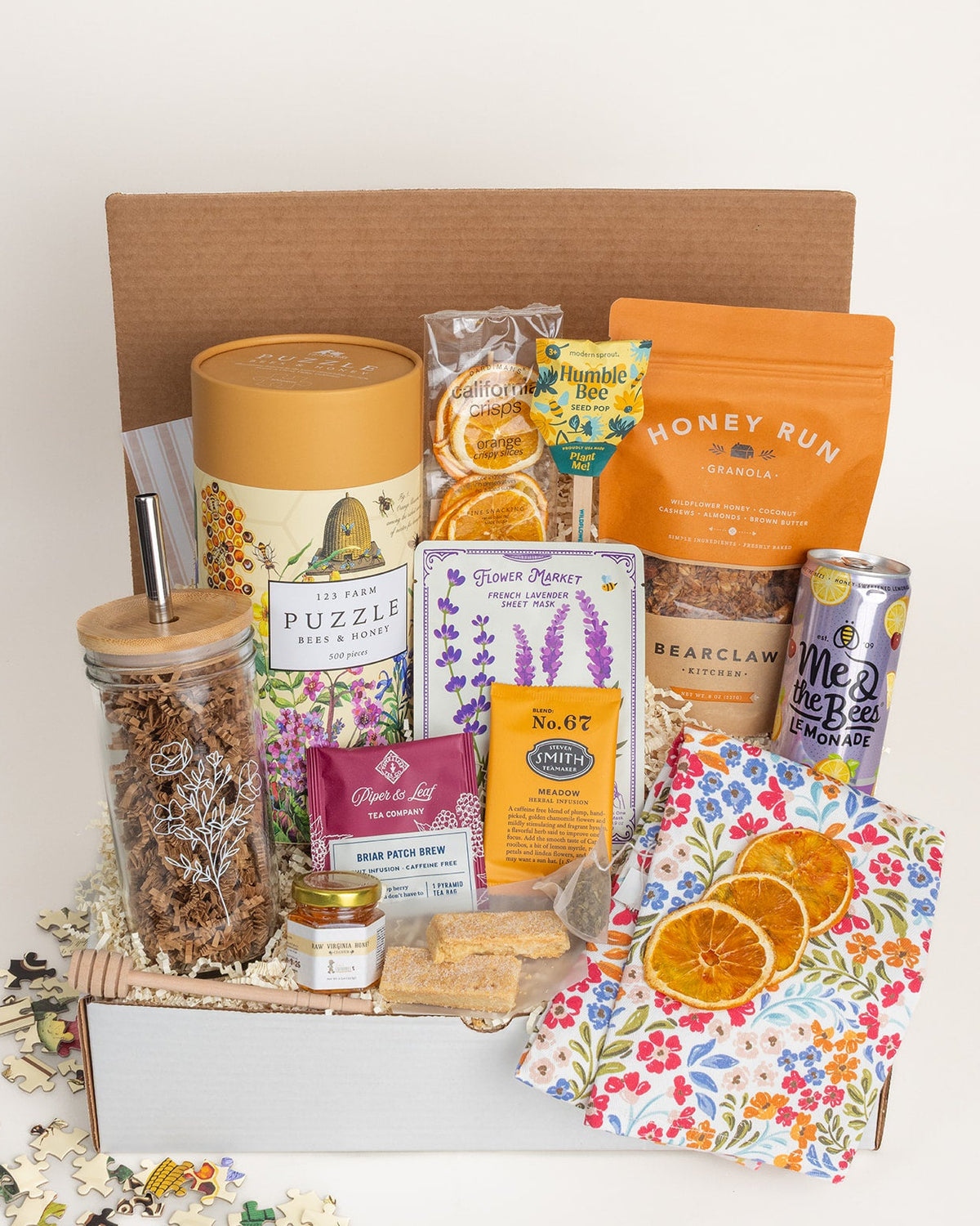 Gift box filled with honey-themed treats, a floral print glass tumbler, tea, granola, a puzzle, and a colorful floral tea towel.