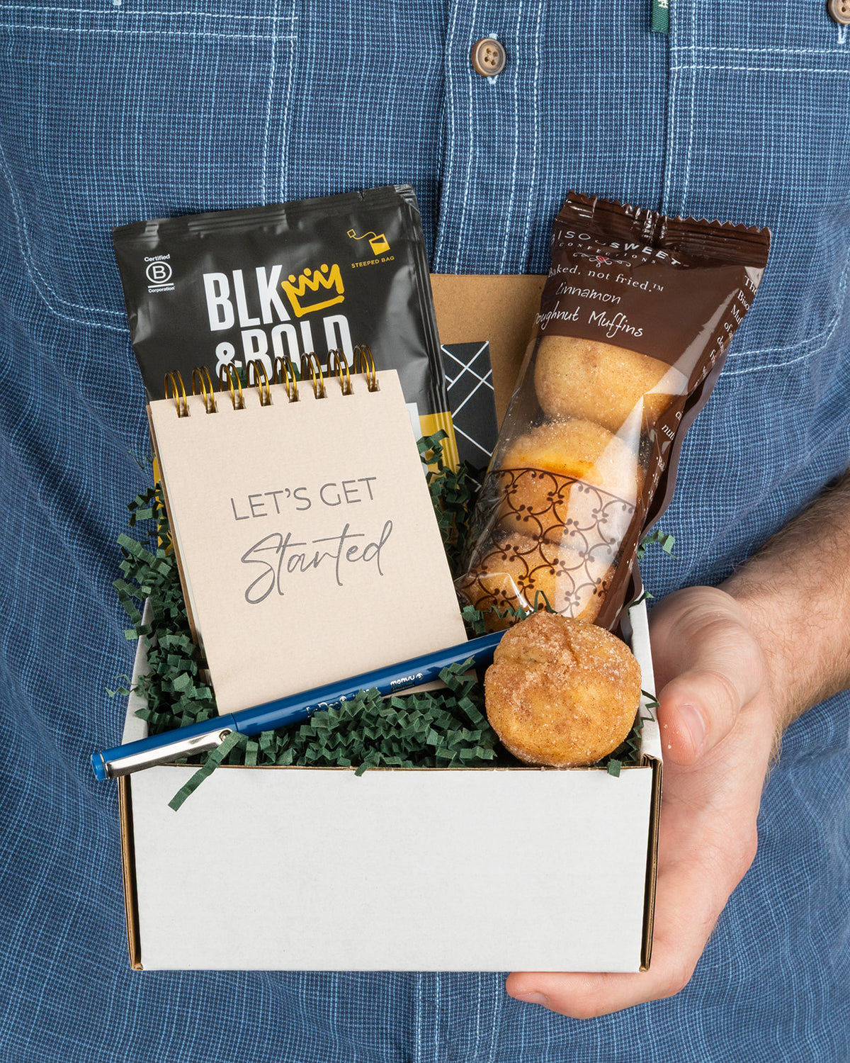 Gift box containing a Let’s Get Started notepad, BLK & Bold coffee, cinnamon donut muffins, and a blue pen on green filler.