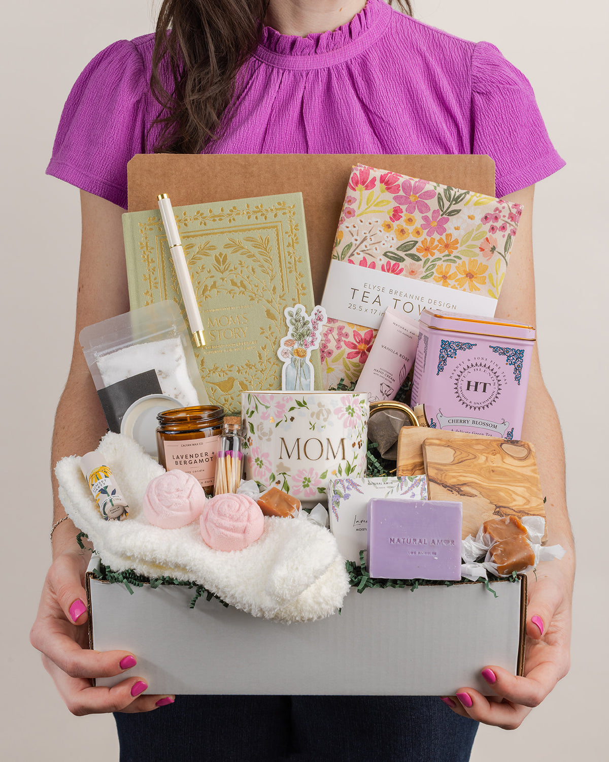 Person holding a gift box filled with a floral Mom mug, keepsake journal, tea, bath salts, candle, and self-care items.