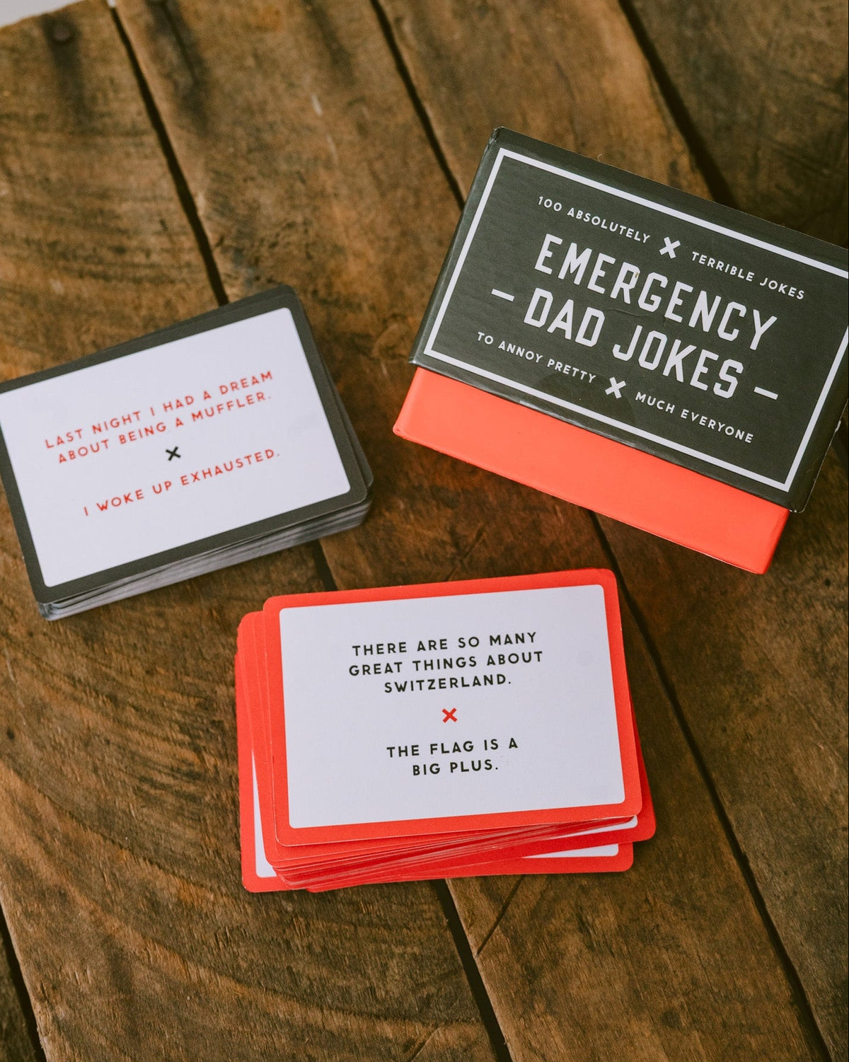 Emergency Dad Jokes card game with black and red box and white joke cards on a rustic wooden surface.