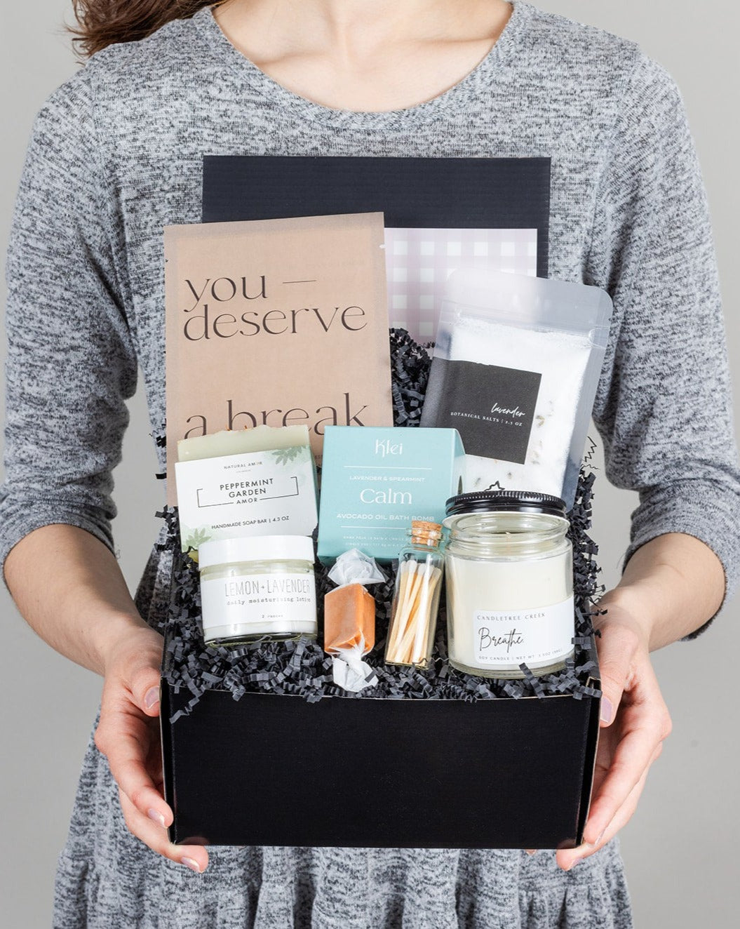 Person holding a black gift box filled with a face mask, candle, matches, soap, lotion, bath salts, and a caramel treat.