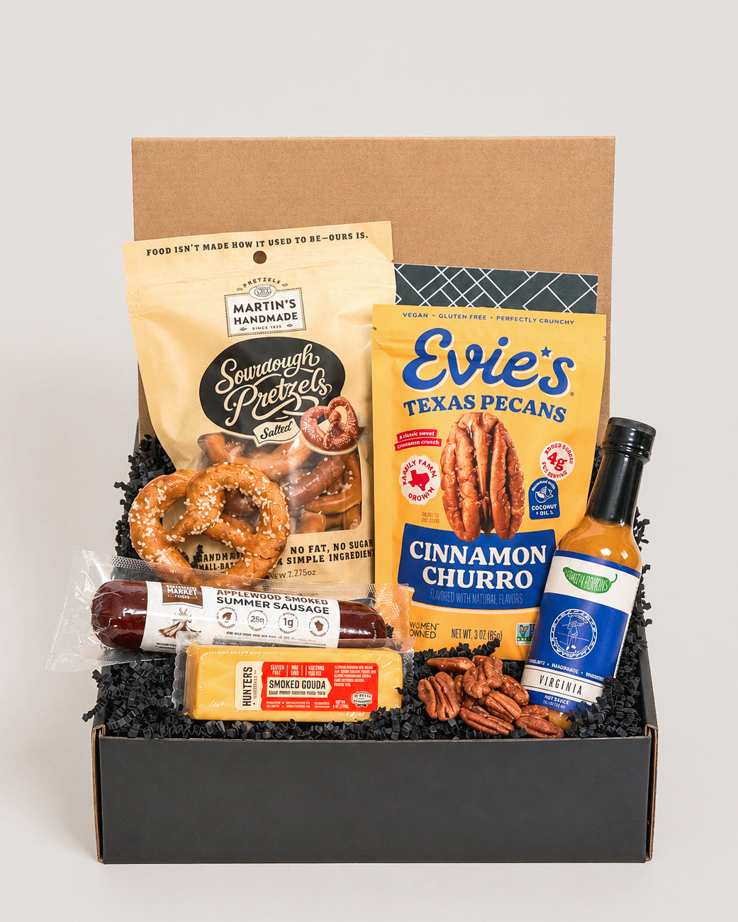 Savory gift box featuring sourdough pretzels, Texas pecans, summer sausage, smoked gouda, and hot sauce on black crinkle paper.