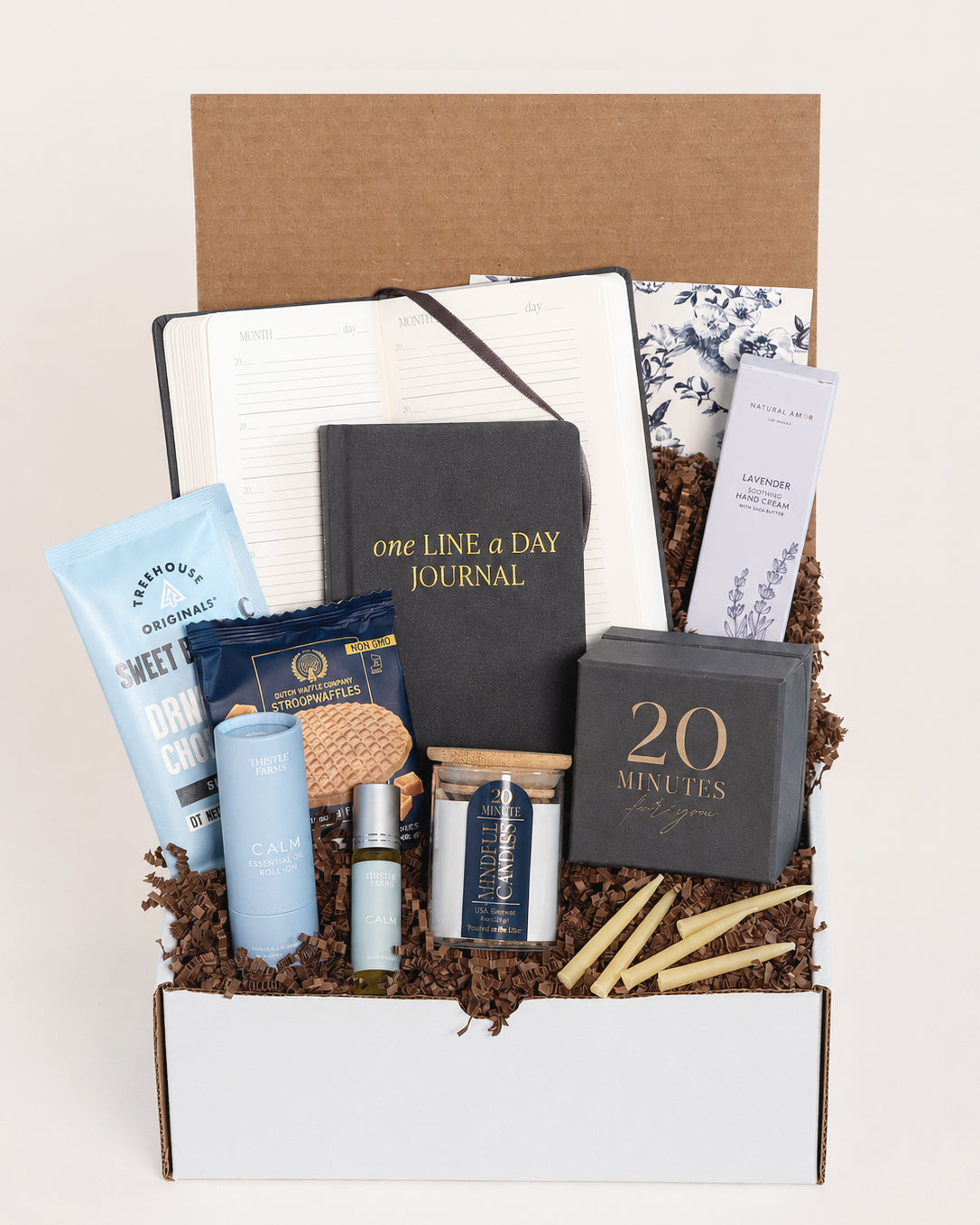 Gift box containing a black journal, beeswax candles, lavender hand cream, stroopwafel cookies, and essential oil roll-on.