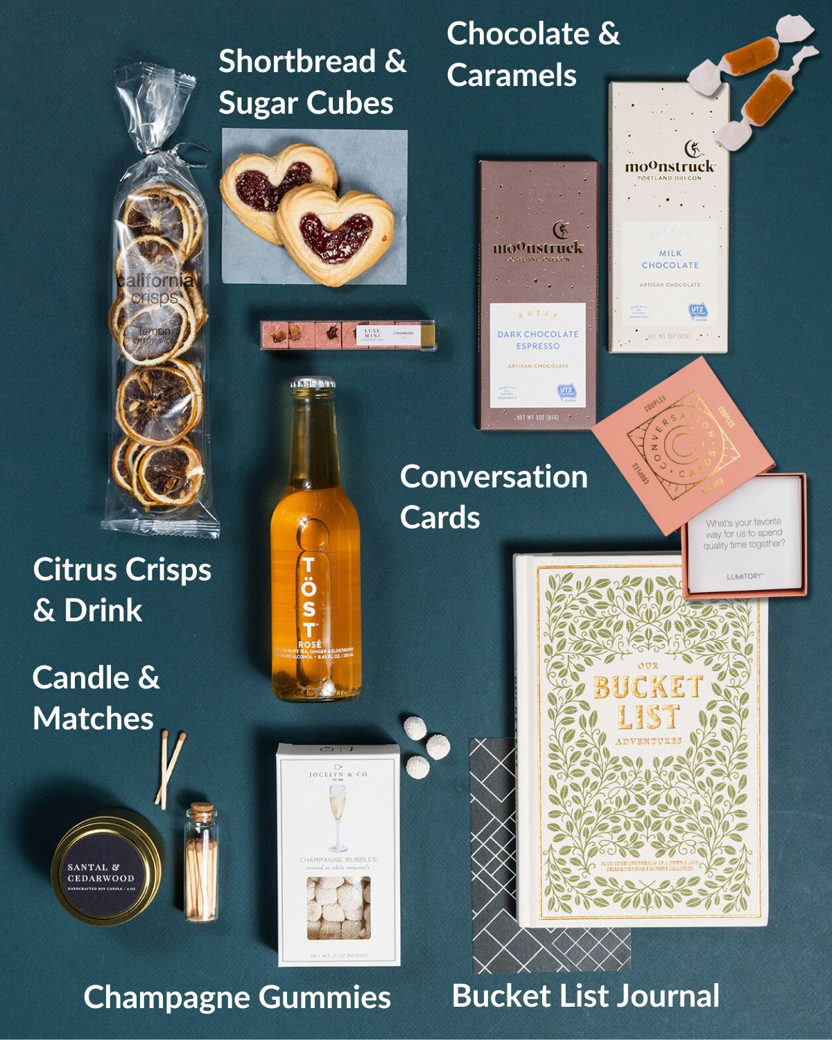 Wedding gift set with Our Bucket List book, conversation cards, TÖST drink, chocolates, heart cookies, and candle on dark teal.