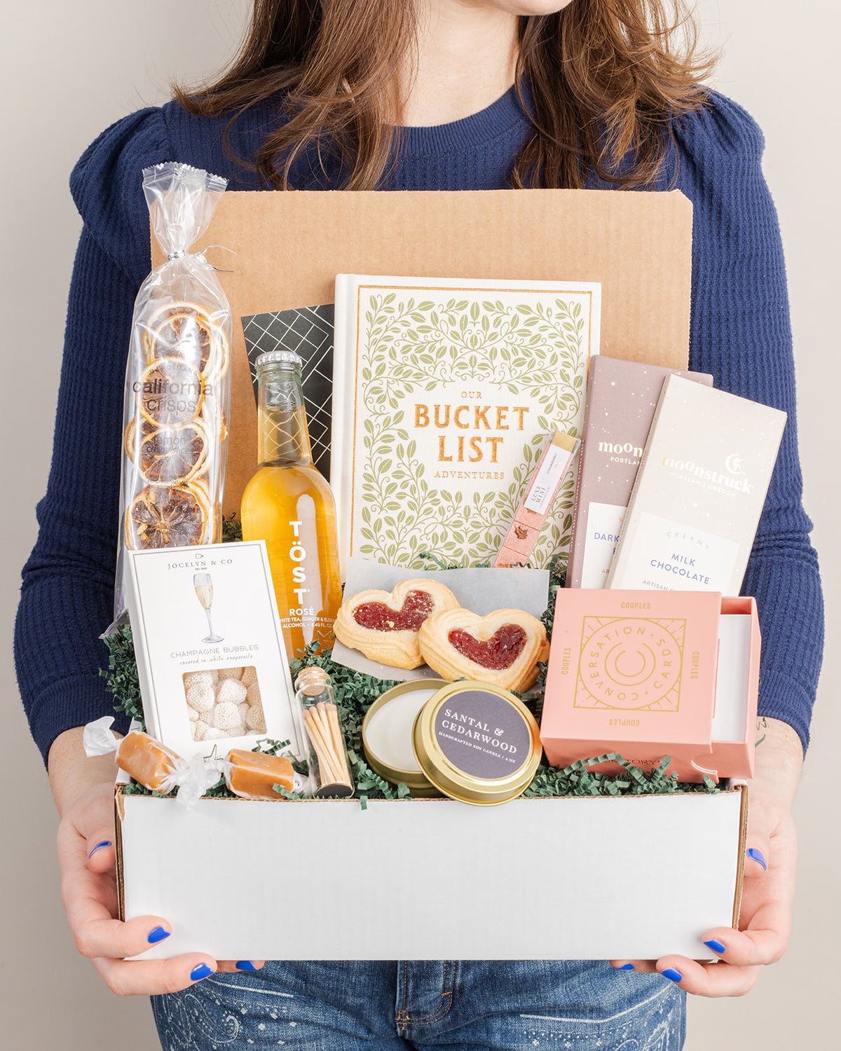 Gift box for couples featuring a bucket list book, conversation cards, sparkling drink, chocolates, heart cookies, and candle.