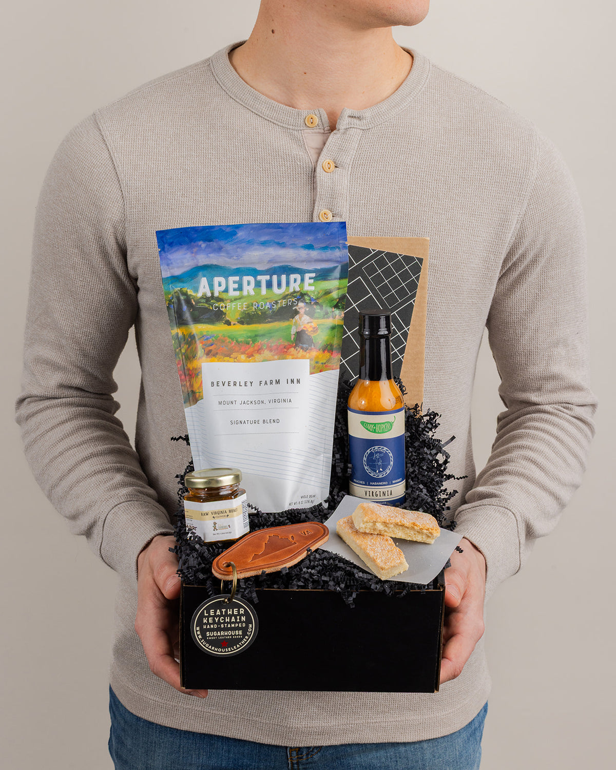 Person holding a black gift box filled with Virginia coffee, hot sauce, honey, shortbread cookies, and a leather keychain.